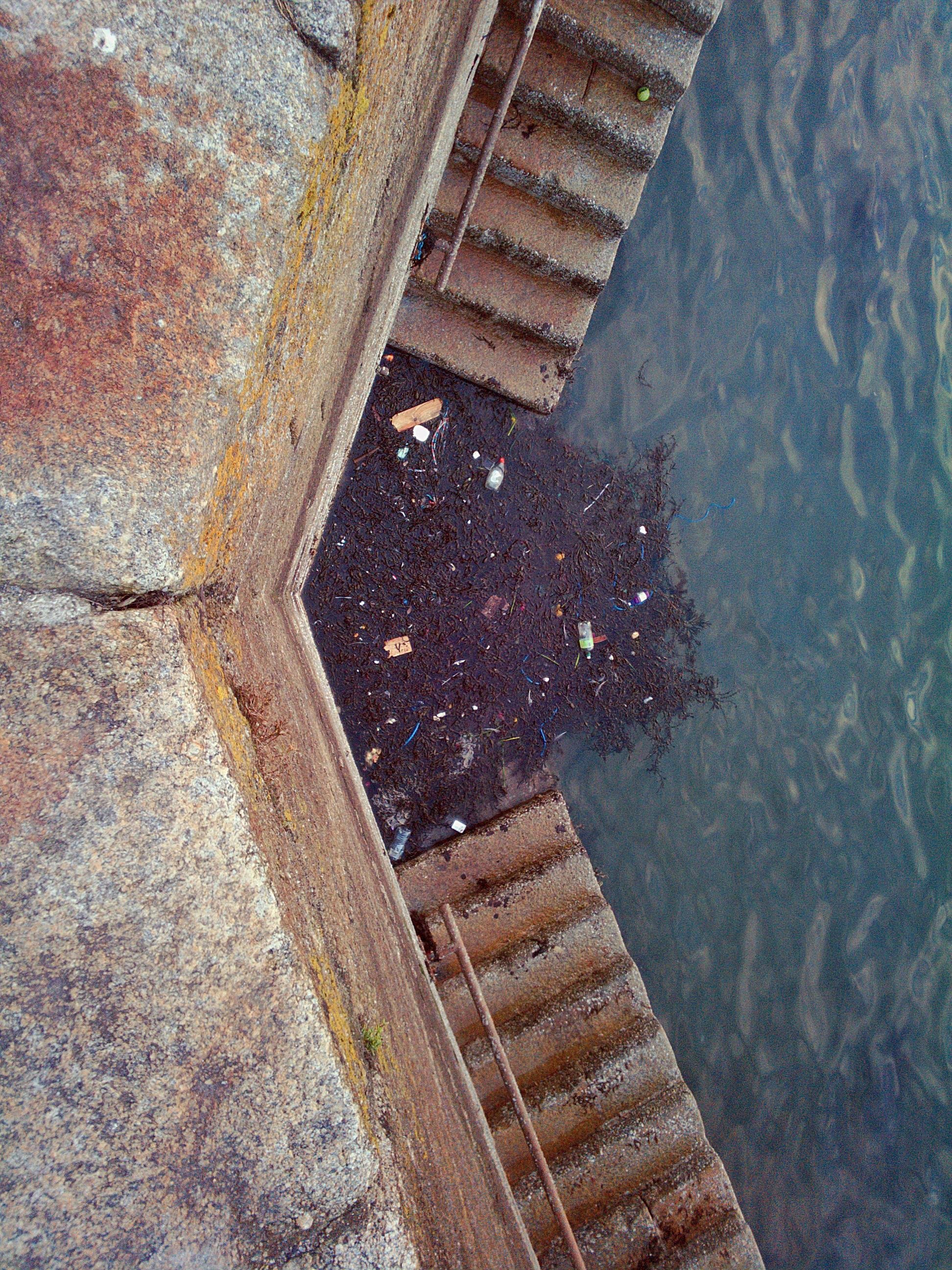 Stone steps lead down to a body of water, with accumulated debris and litter on the steps and along the water's edge. The water is slightly rippled and reflects a muted color.