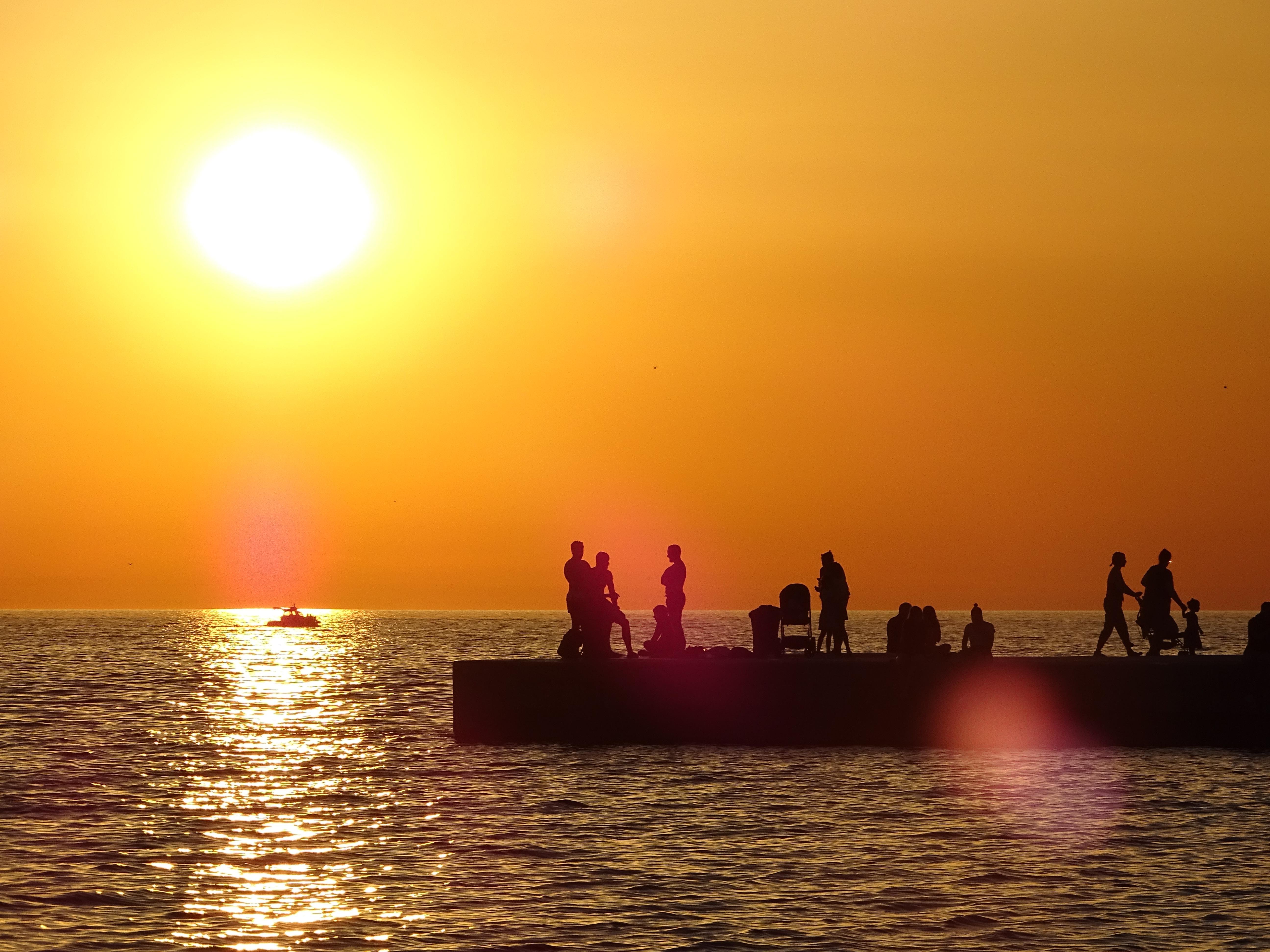 A vivid sunset casts a golden hue over a calm sea with the sun positioned prominently in the upper left quadrant. Silhouetted figures of various people are visible on a pier.