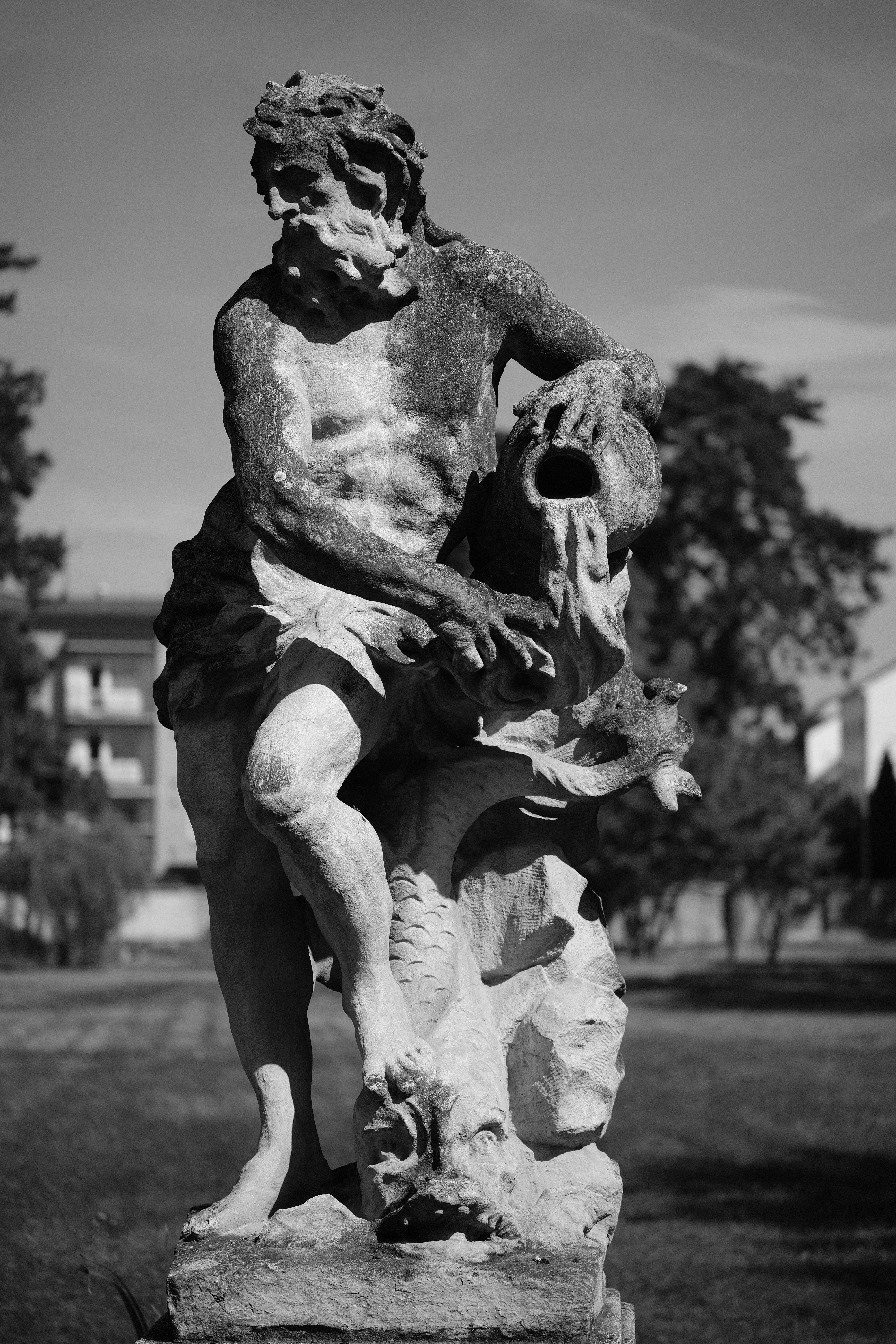 Black and white photo of a weathered stone statue depicting a muscular figure with an expressive face, wild curly hair, and partial drapery around the waist. The figure is holding up a pot with one hand, and the other is placed on a mythical creature's head, possibly a dragon, featuring sharply detailed scales (not sure tbh, I'd the part where the statue's second hand meets the creature is pretty busy). The background shows blurry trees and a building.
