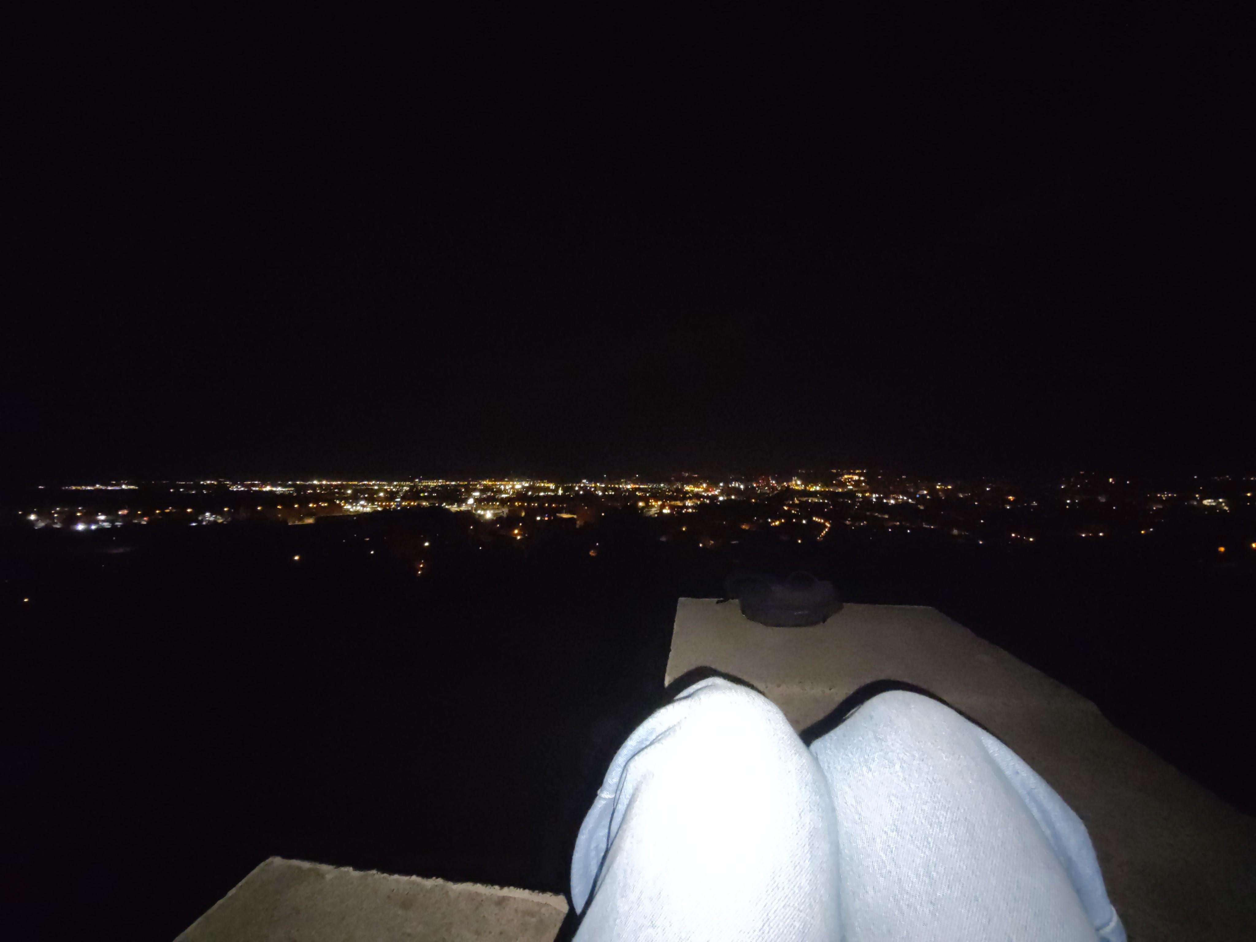 Nighttime view over a cityscape from a high vantage point, featuring scattered city lights stretching into the distance. Foreground shows my legs, knees bent, with feet resting on a concrete ledge. The legs are covered with blue jeans. The phone's LED flash overexposed them, so they seem very light.